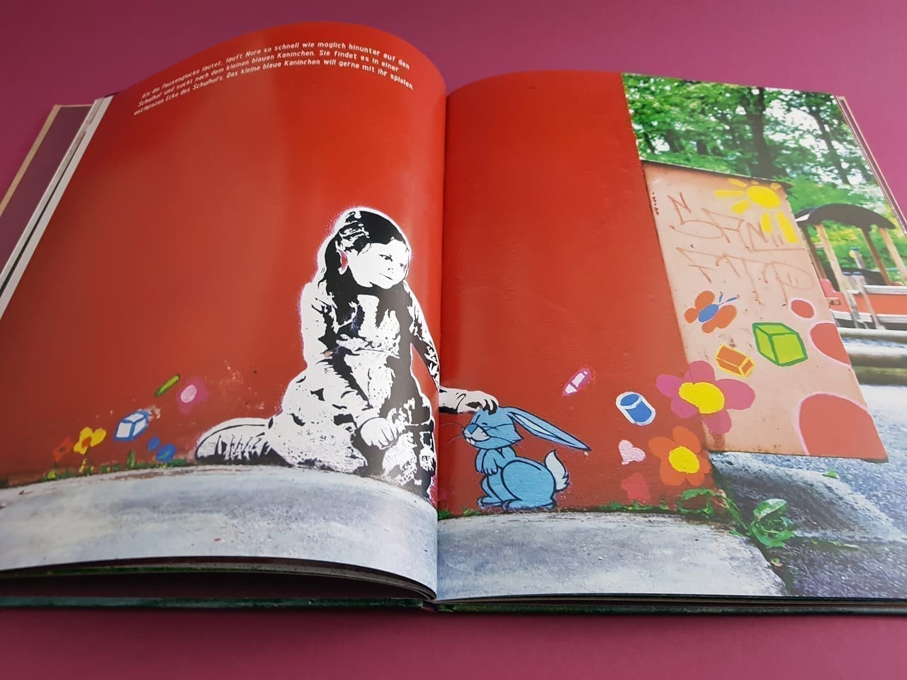 Im aufgeschlagenen Buch „Nora und das kleine blaue Kaninchen“ von Gingko Press sieht man das Bild einer roten Fläche mit der schwarz-weißen Zeichnung eines kleinen Mädchens, kniend und ein kleines blaues Kaninchen streichelnd. Um sie herum sind einige bunte Blumen gezeichnet. Über ihrem Kopf ist ein kleiner Text in weißer Schrift zu lesen. Wahrscheinlich befindet sich diese rote Wand an einem Spielplatz: auf der rechten Buchseite sieht man im Hintergrund Bäume und Spielplatzgeräte.