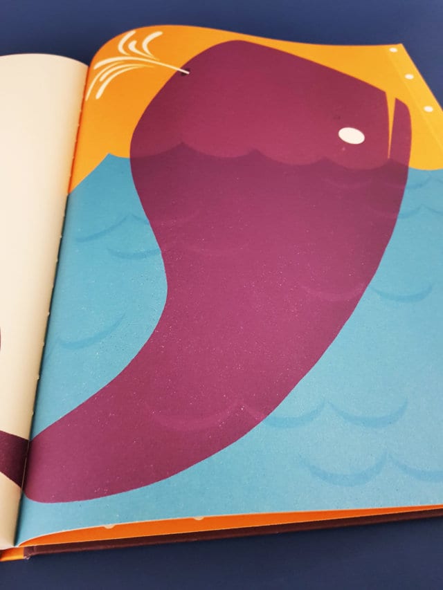 Aufgeschlagene rechte Seite des Kinderbuchs ,Alle meine Tiere‘ von Hendrik Hellige mit dem Bild eines magentafarbenen Wals. Der Wal befindet sich im Wasser, wobei sein Kopf aus dem Wasser herausschaut. Das Wasser ist blau, der Hintergrund über dem Wasser ist gelb.