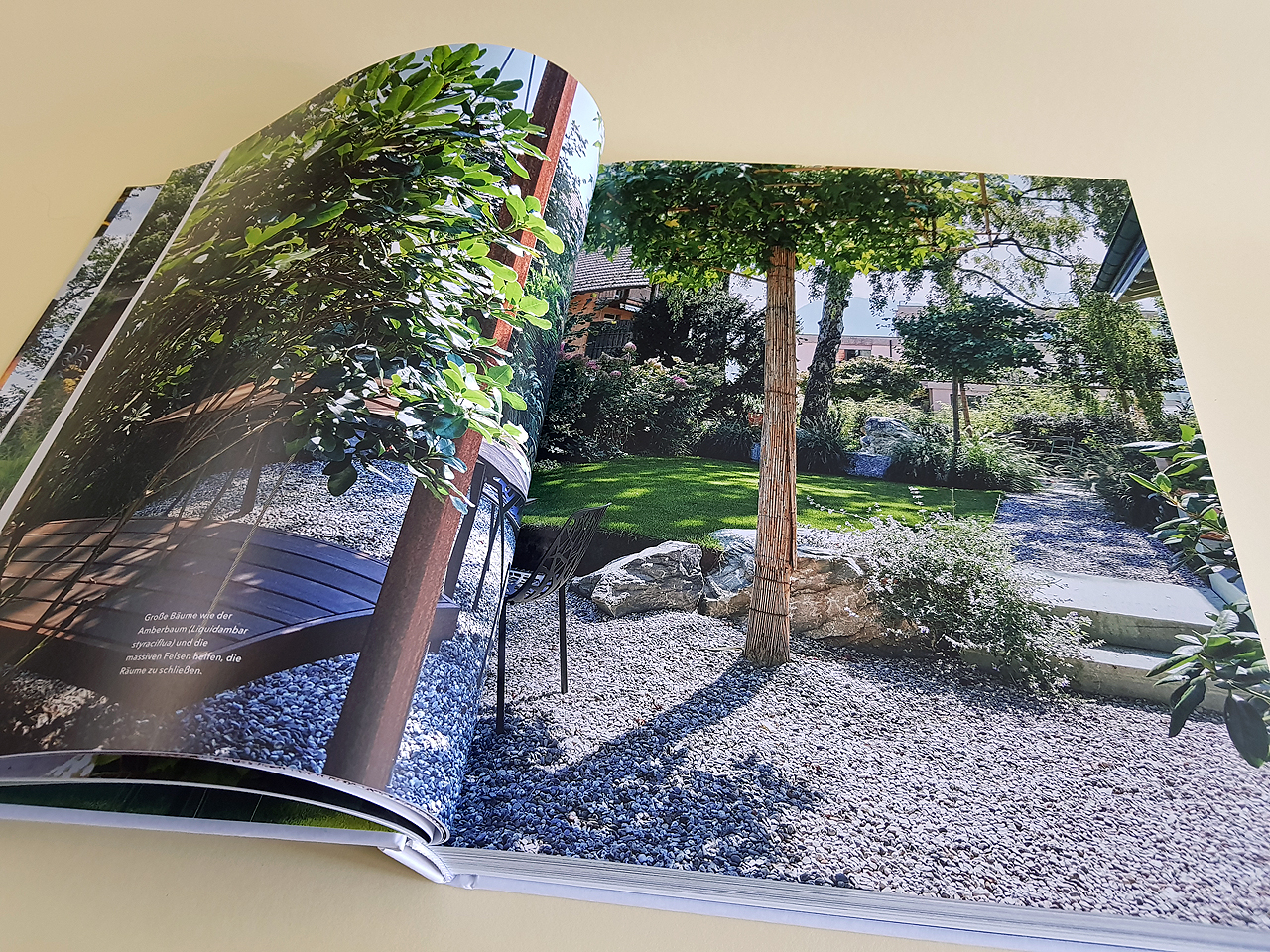 Aufgeschlagene Doppelseite des Buches „Gärten des Jahres 2022“. Zu sehen ist ein großer Garten mit jeder Menge Bäumen und Pflanzen sowie einer Holzbank links im Bild. Auf der rechten Seite sieht man einen Weg mit weißen Schottersteinen.