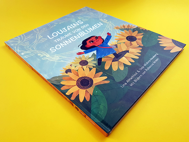 Schräg liegendes Cover des Kinderbuchs ‚Loujains Träume von den Sonnenblumen‘ von Lina Al-Hathloul und Uma Mishra-Newbery aus dem Verlag minedition. Vor hellblauem Himmel steht Loujain, ein junges Mädchen, auf einem Sonnenblumenfeld. Sie steht auf einer Sonnenblume in einem blauen Kleid und hat die Arme ausgebreitet. Über ihrem Kopf ist der Titel zu lesen.